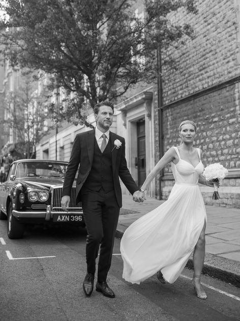 A bride in a flowing white gown and a groom in a dark suit walk hand in hand down a city street, with a classic car parked nearby and a stone building in the background.