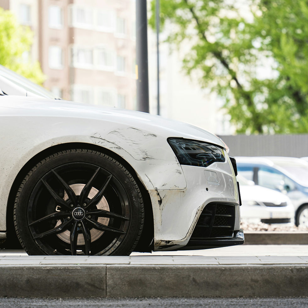 A white Audi car with noticeable scratches and scuffs on the front bumper and fender is parked outdoors. The black alloy wheels contrast with the car's body, and a blurred background of trees and other vehicles is visible.