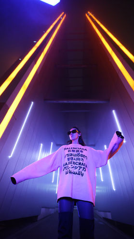 A woman wearing a large Balenciaga jumper and sunglasses stands in a futuristic setting with bright neon lights, including orange and blue hues, creating a dramatic atmosphere.