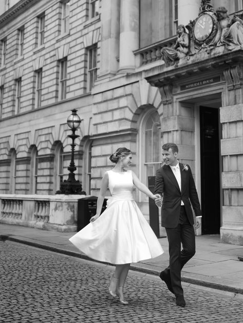 A couple joyfully walks hand in hand on a cobblestone street, with the woman twirling in a knee-length white dress and the man in a dark suit, set against the backdrop of an elegant, historic building.