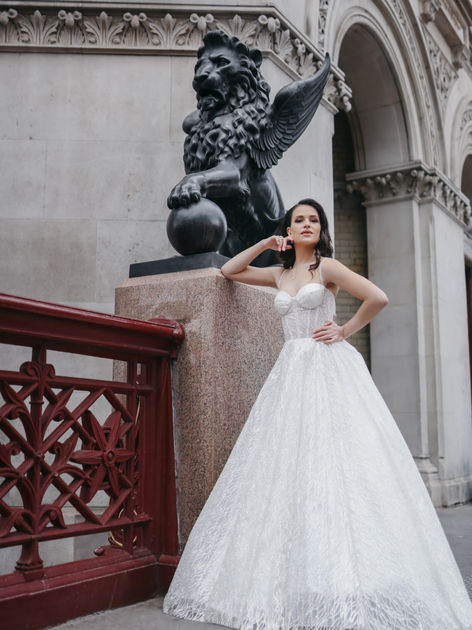 A woman in an elegant white wedding dress poses confidently beside a large winged lion statue on a stone pedestal. She stands on a bridge with intricate red railing, set against a backdrop of ornate stone architecture.