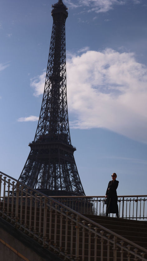 A woman in a long coat and hat stands on the stairs, with the Eiffel Tower standing out against a partially cloudy sky in the background.