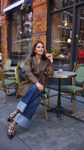 A woman sits at an outdoor café table, dressed in a stylish brown blazer and wide-legged jeans with rolled cuffs. She wears studded black heels and holds a black handbag, while a small coffee cup rests on the table beside her. The café has a brick facade and green wicker chairs, creating a cozy, urban setting.