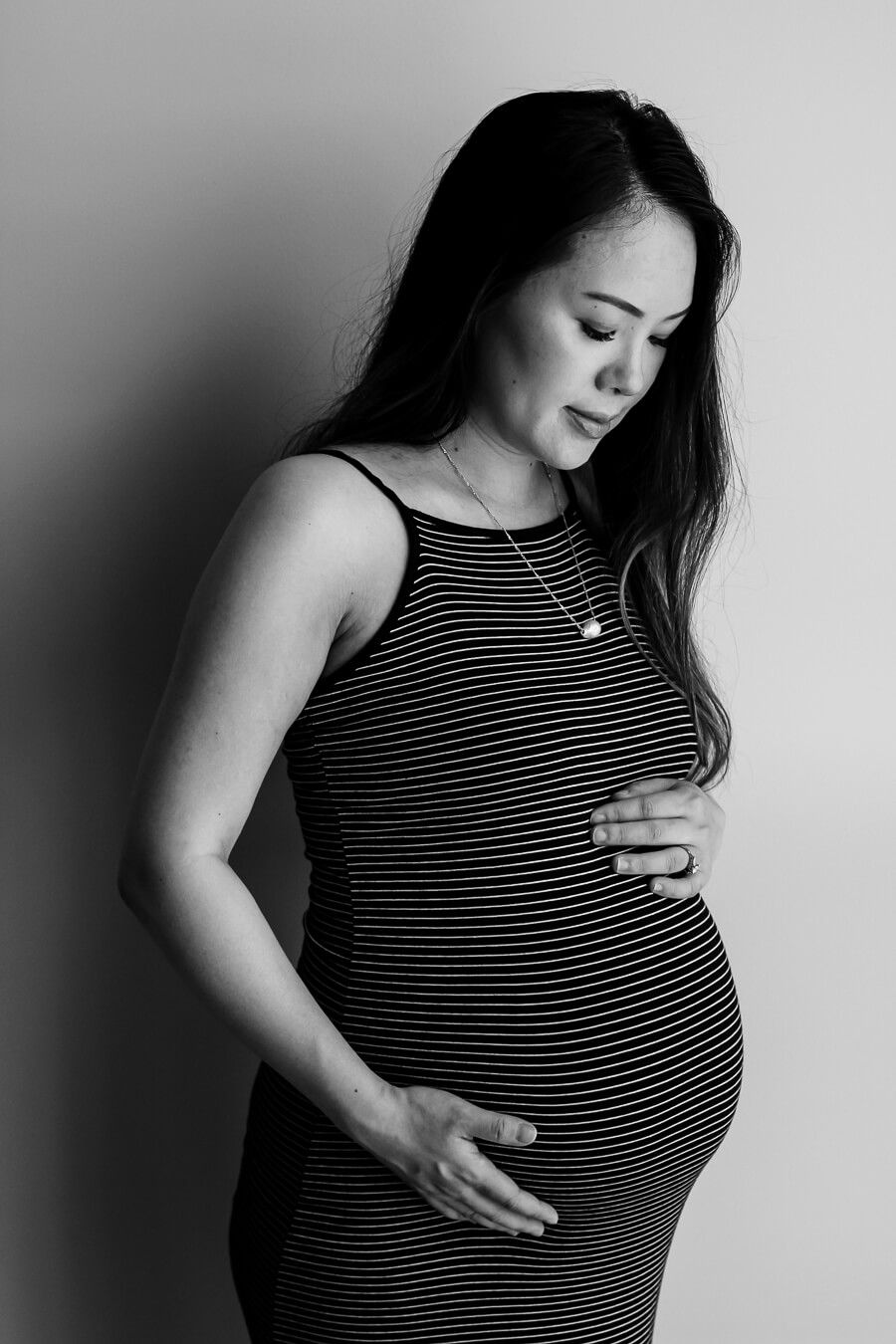 A black and white photo of a mother-to-be standing and looking down at her bump. She is wearing a dark colored dress with thin white stripes.