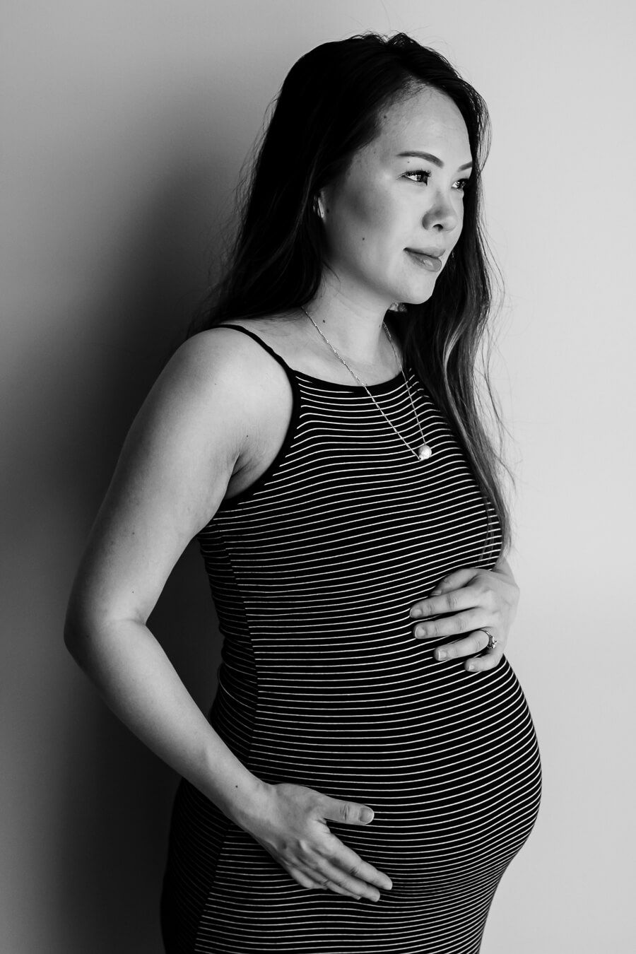 A black and white maternity portrait of a pregnant mama-to-be with long hair. She is wearing a dark dress with thin white stripes.