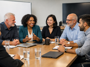 Eye-level view of a modern conference room where a diverse group of professionals sits around a wooden table in active discussion. An African American woman in a teal blazer speaks while colleagues of different ages and ethnic backgrounds listen, smile, and engage. Laptops, notebooks, coffee mugs, and glasses of water are arranged across the table, creating a collaborative, professional atmosphere.