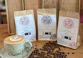 Latte with three coffee bags and scattered beans on wooden table.