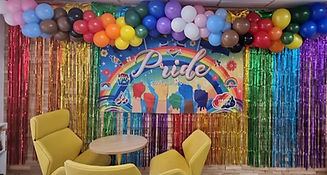 Colorful balloons above a rainbow Pride banner with gold fringe; Pride ofure events.