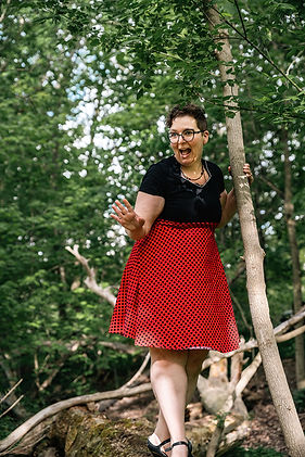 A woman with dark curly hair is standing with one hand on a tree and her other hand reaching out. She is wearing a knee length black dress with a bottom half that is red with black polka dots. She has an excited expression - like she is saying something very passionately. 