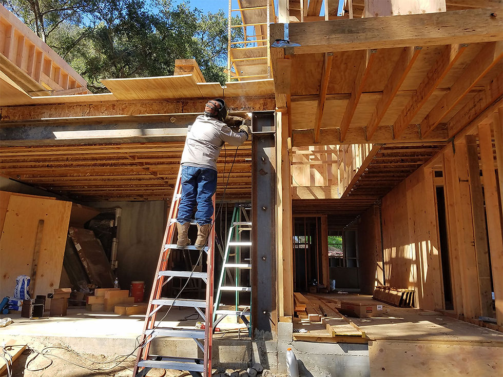 Worker on ladder with helmet, welding on steel construction. Sunlit open frame, scattered materials, and trees in background. Rustic setting.
