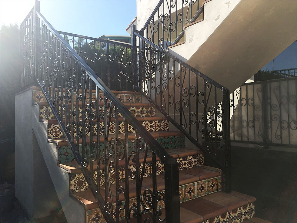 Ornate tiled staircase with decorative black wrought iron railings, set outdoors under sunlight, casting shadows on a wall.