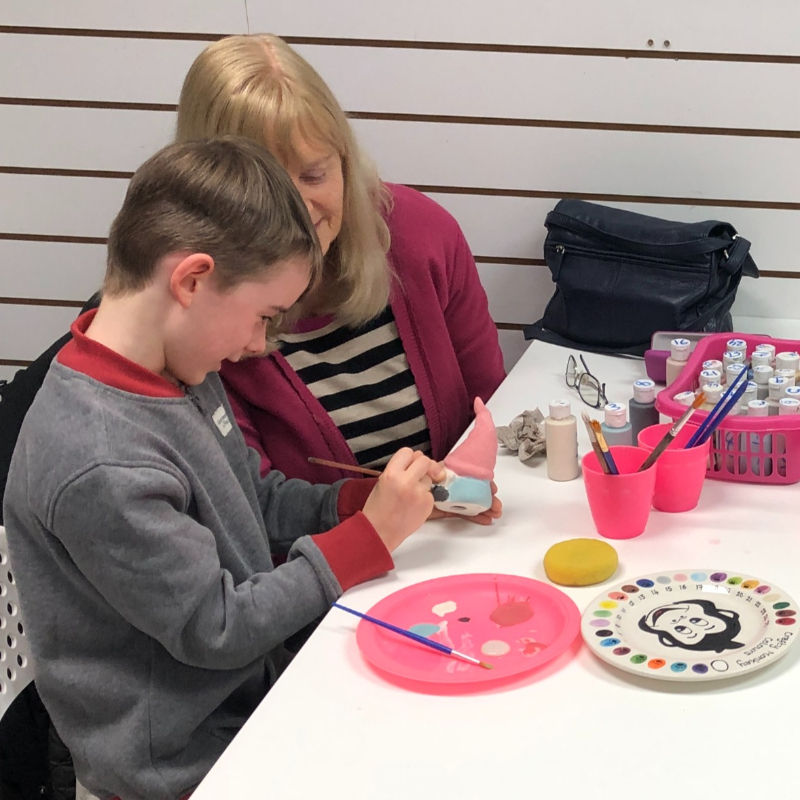 A grandmother spending quality time with her grandchild while he paints a gnome at a pottery painting studio