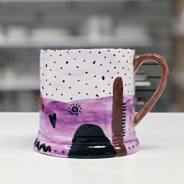 Desert theme painted on a mug in purple hues with Crafty Monkey Pottery Painting Studio in the background.