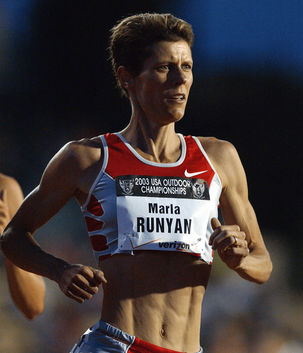 Marla Runyan, atleta norte-americana legalmente cega devido à Doença de Stargardt, aparece competindo no Campeonato Outdoor dos EUA de 2003. Ela veste uniforme vermelho e branco com número de peito identificando '2003 USA Outdoor Championships – Marla Runyan – Verizon'. A imagem transmite esforço e determinação, destacando sua trajetória como primeira atleta com deficiência visual a participar dos Jogos Olímpicos e múltipla medalhista paralímpica.