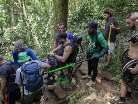Grupo de homens auxilia um cadeirante em cadeira adaptada durante percurso em trilha na mata, superando trecho com pedras e desníveis em meio à vegetação densa.