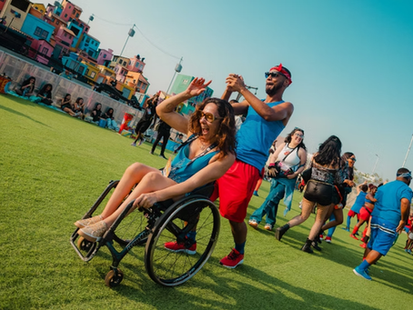 Mulher usando cadeira de rodas participa de um momento de lazer em um espaço aberto com gramado. Um homem a empurra enquanto os dois parecem se divertir e dançar. Ao redor, várias pessoas caminham e interagem em um ambiente festivo, com casas coloridas ao fundo e céu azul.