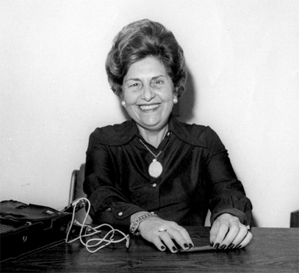 Dorina de Gouvêa Nowill, educadora e ativista brasileira pela inclusão das pessoas cegas, aparece sorridente em fotografia em preto e branco. Ela está sentada à mesa, usando blusa escura e colar com pingente grande, segurando um pequeno objeto na mão. À frente, há uma bolsa ou maleta com fios conectados, sugerindo um ambiente de trabalho ou entrevista. A imagem transmite simpatia e determinação, refletindo sua papel histórico como fundadora da Fundação Dorina Nowill e pioneira na difusão do sistema Braille no Brasil