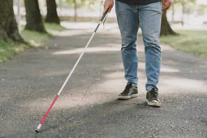 Pessoa caminhando em uma estrada arborizada, utilizando uma bengala branca com faixas vermelhas, que é o símbolo de acessibilidade para pessoas com surdocegueira. A bengala está sendo usada para explorar o chão e auxiliar na locomoção. A pessoa veste calça jeans azul, tênis pretos e uma camiseta escura. O ambiente é um caminho pavimentado em meio a árvores, sugerindo um espaço ao ar livre, como um parque.