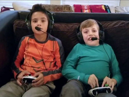Dois meninos, de aproximadamente 10 anos, sentados lado a lado em um sofá escuro, jogando videogame. O menino à esquerda veste uma camiseta laranja e cinza, enquanto o da direita usa uma camiseta verde. Ambos utilizam headsets pretos com microfones e seguram controles de videogame brancos. Eles olham para a frente com expressões de concentração. Ao fundo, vê-se parte de uma sala com almofadas e uma manta colorida.