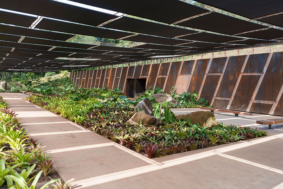 Fotografia da área externa da Galeria Cosmococa no Instituto Inhotim. A imagem mostra um pátio coberto por uma estrutura metálica de brises pretos retangulares, que criam um jogo geométrico de luz e sombra no chão de terra batida. À direita, uma longa parede de aço corten, com tom ferrugem característico, possui uma abertura central. No centro, jardins internos com bromélias verdes e avermelhadas, folhagens tropicais e grandes rochas decorativas compõem o paisagismo. O ambiente transmite uma sensação de refúgio e integração entre arquitetura brutalista e natureza.