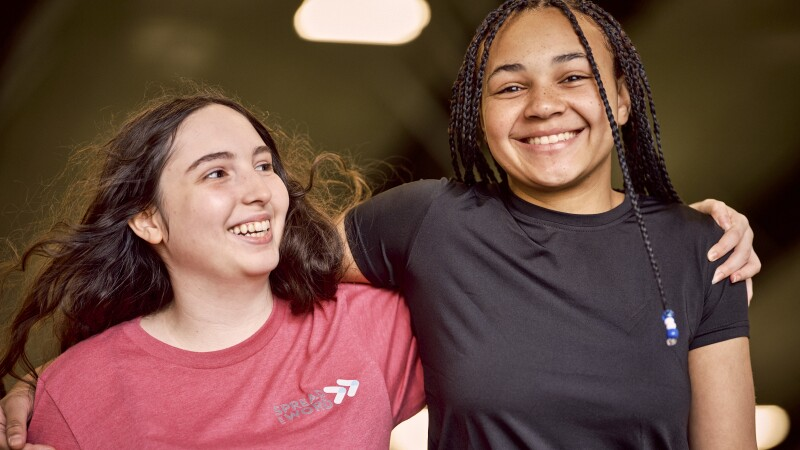 Duas jovens sorridentes posam abraçadas pelos ombros em um ambiente interno com iluminação quente. À esquerda, uma jovem branca com longos cabelos castanhos ondulados veste uma camiseta rosa com um pequeno logotipo branco. À direita, uma jovem negra com o cabelo em tranças longas e finas (box braids), adornadas com algumas contas azuis e brancas na ponta, veste uma camiseta preta. Ambas demonstram uma expressão de alegria e cumplicidade. O fundo está desfocado, destacando a amizade entre as duas.