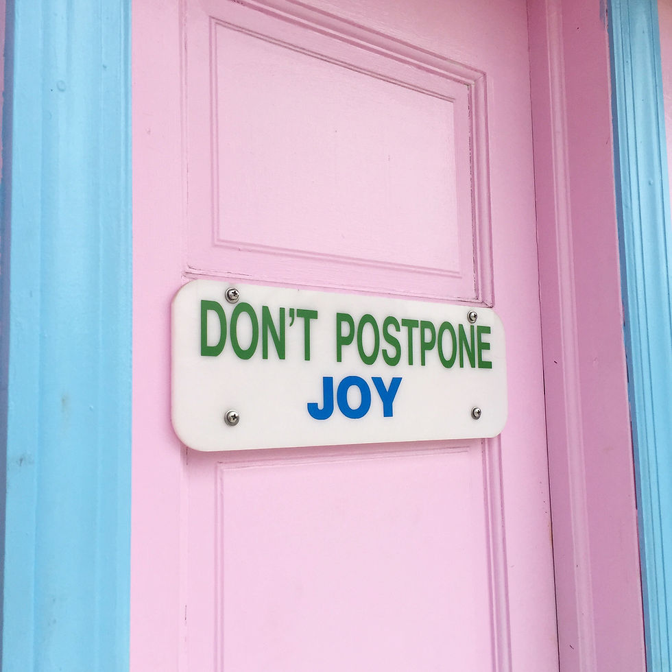 A sign which reads DON’T POSTPONE JOY attached to a bubblegum pink door
