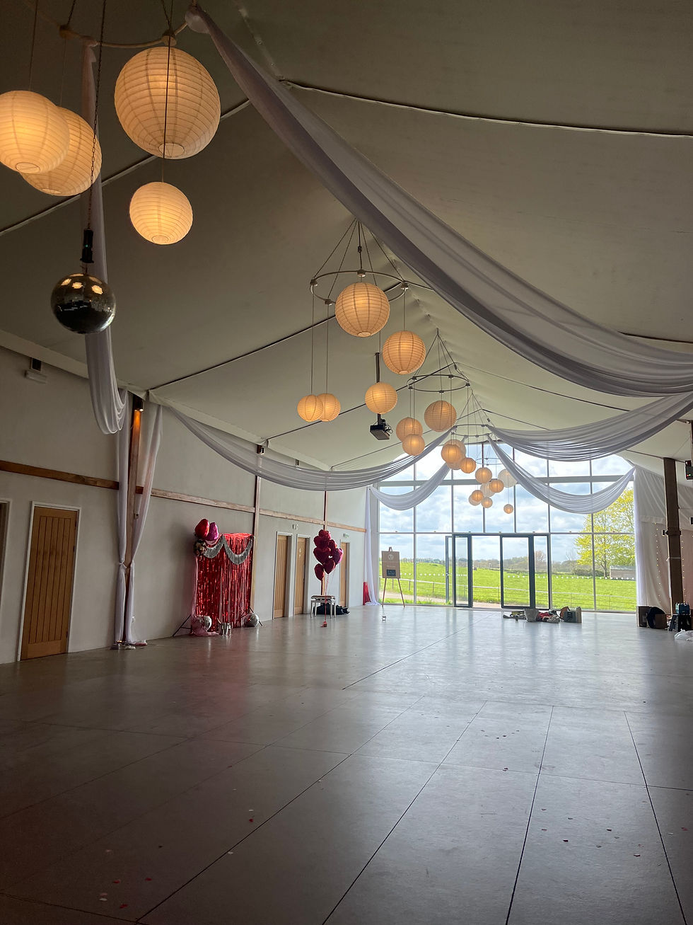 Empty decorated hall with hanging lanterns, white drapes, red balloon display, and floor-to-ceiling windows overlooking green fields.