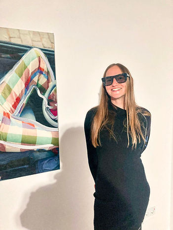 Smiling woman in black with sunglasses stands beside colorful plaid artwork in a sparse room, conveying a cheerful mood.