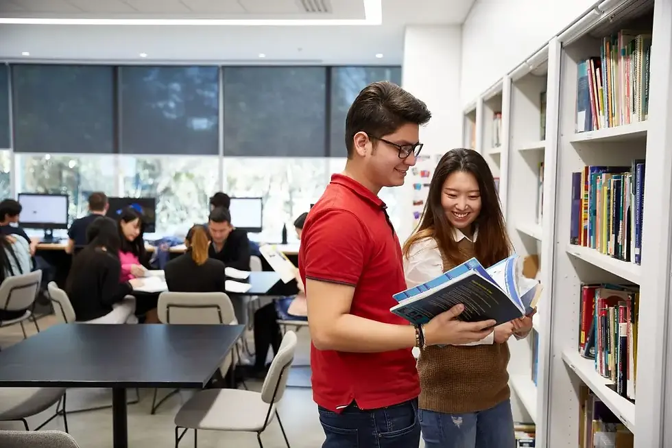 Two students studying together in a library, reviewing textbooks beside bookshelves in an academic setting.