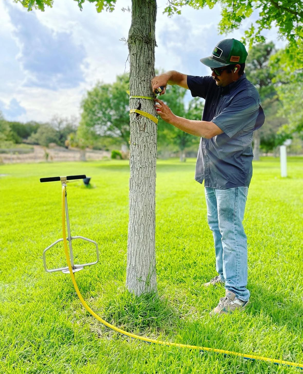 Tejas Pest and Weed Control tech measuring a tree trunk beside a deep-root soil injection probe for Odessa TX tree treatments