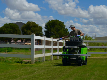 Tejas Pest and Weed Control tech on a lime-green Steel Green stand-on sprayer with handheld wand along a white fence on a lush West Texas lawn