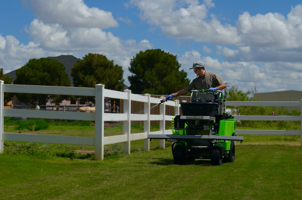 Tejas Pest and Weed Control tech on a lime-green Steel Green stand-on sprayer with handheld wand along a white fence on a lush West Texas lawn