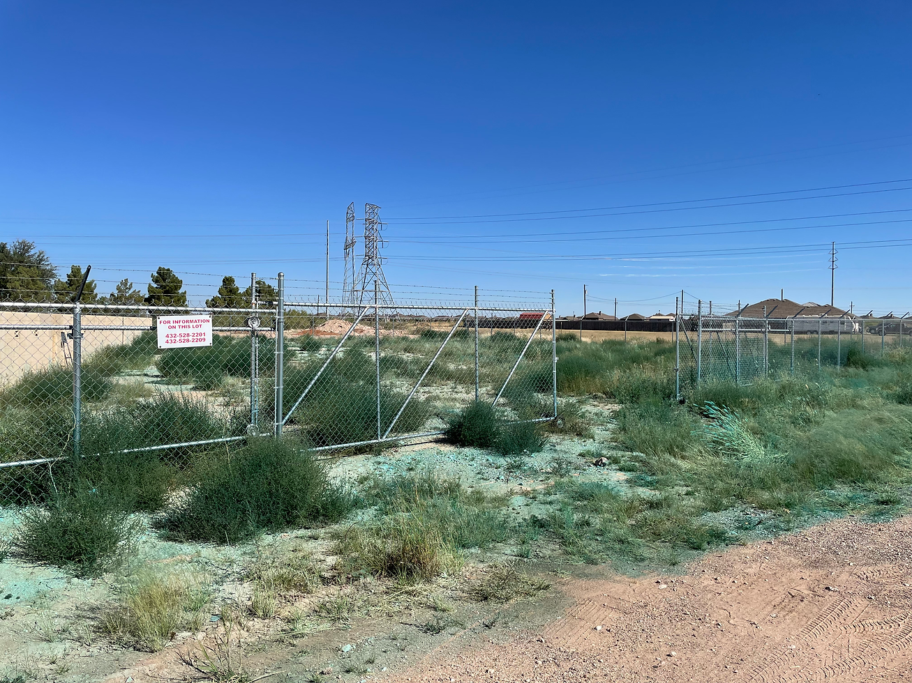 Chain-link fence line and open lot with clumped West Texas weeds and teal marker dye from a Tejas Pest and Weed Control treatment. Fence-line weed control for Andrews and Seminole, Texas.