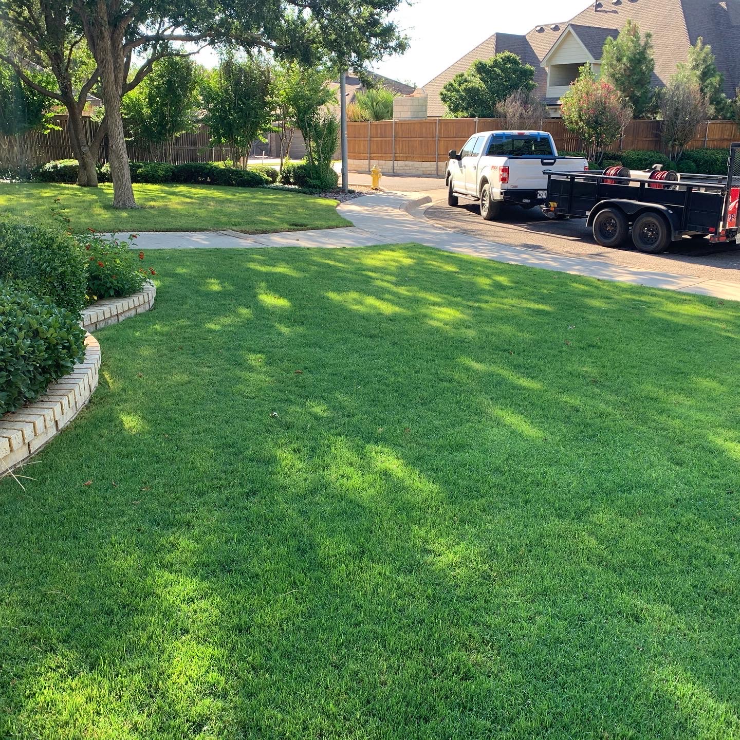 Tejas Pest and Weed Control white Ford pickup and black spray trailer on a lush green Midland residential lawn beside a brick garden bed and privacy fence