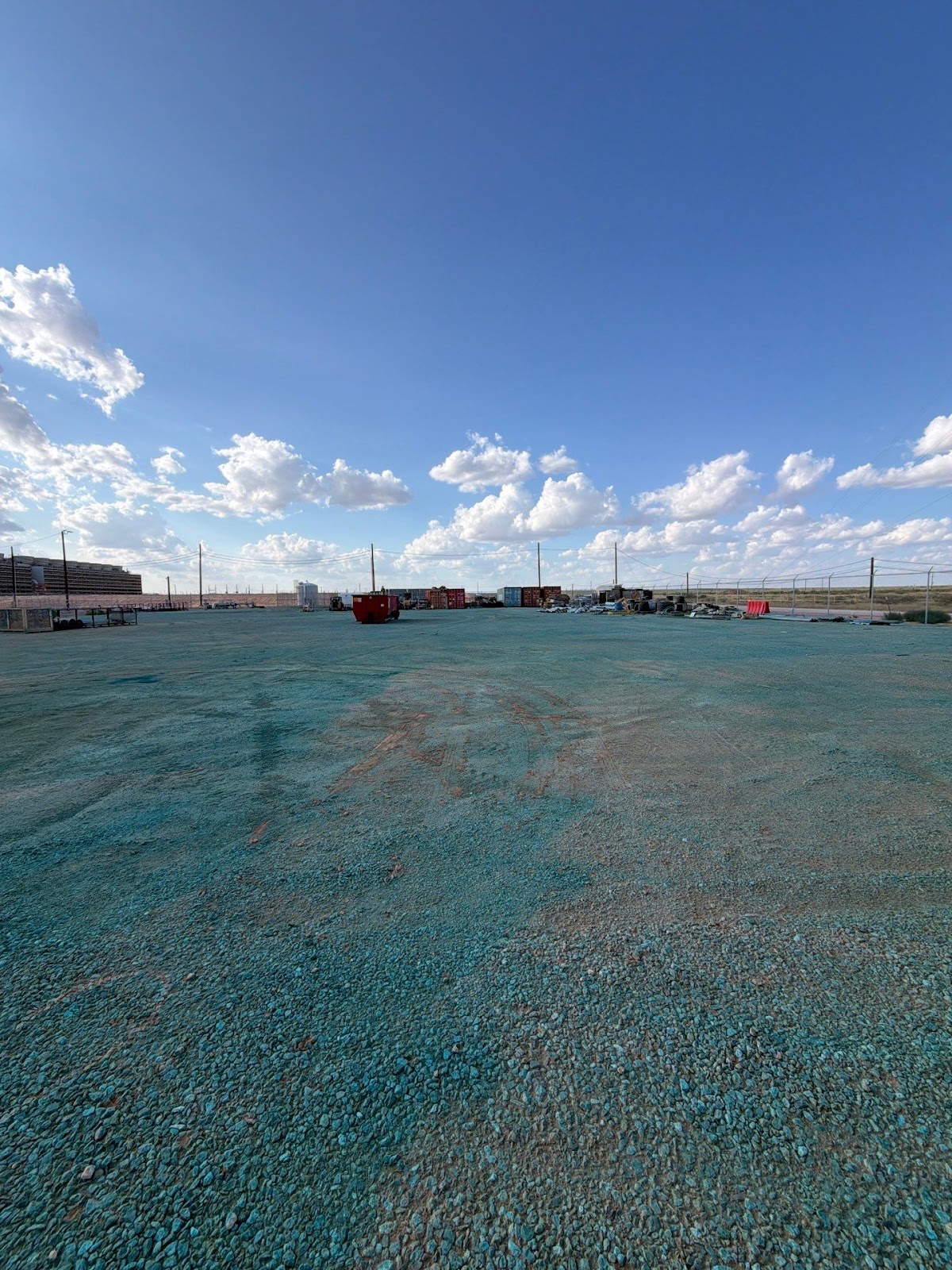Wide industrial lot with teal herbicide indicator dye on gravel; shipping containers and chain-link fence under West Texas sky