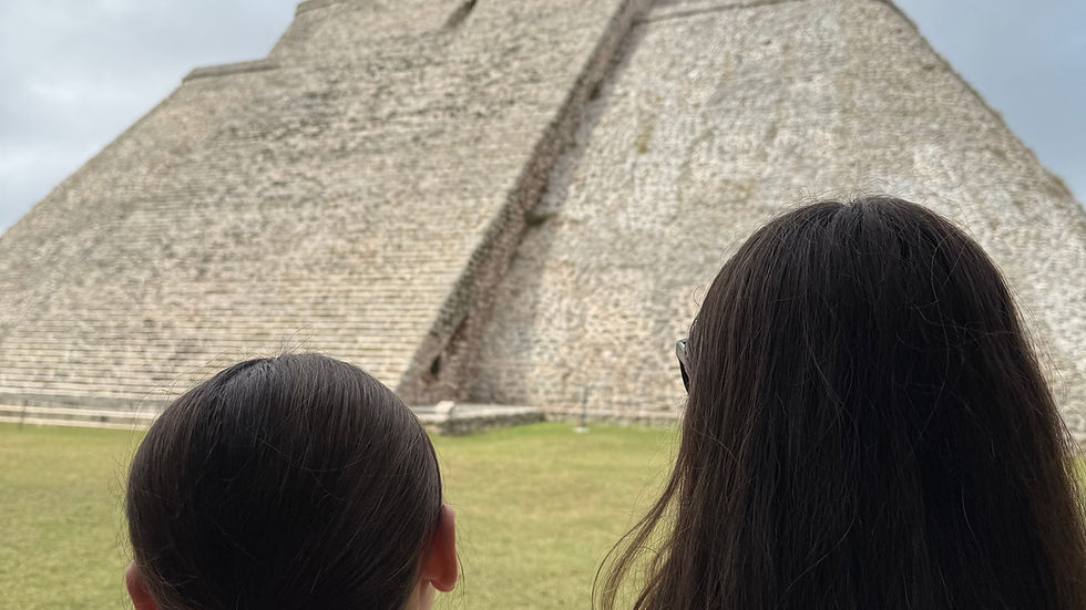 Two people with long hair, seen from behind, stand before a large ancient stone pyramid under a cloudy sky, evoking a sense of awe.