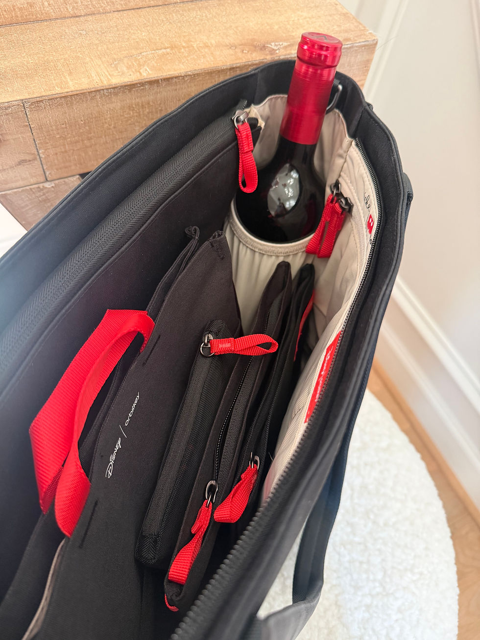 Open black wine tote with red straps holds a bottle of red wine and folders; Disney text is visible.