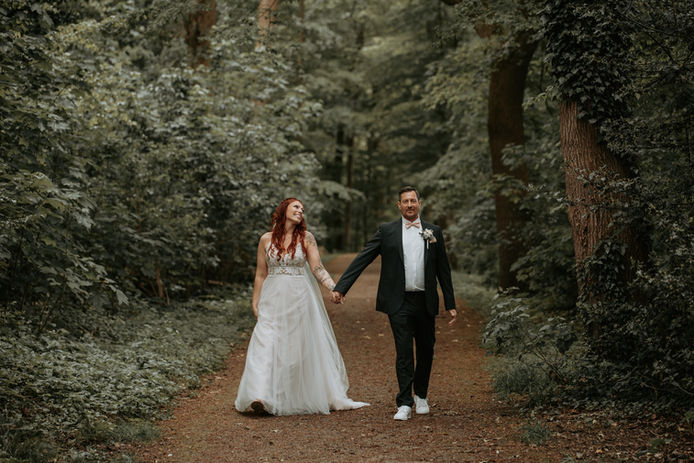Ein Hochzeitspaar spaziert Hand in Hand einen idyllischen Waldweg entlang. Die Braut mit roten Haaren und einem fließenden weißen Kleid schaut ihren Bräutigam liebevoll an, während er entspannt nach vorne blickt. Ein natürliches, romantisches Hochzeitsfoto voller Leichtigkeit und Verbundenheit.