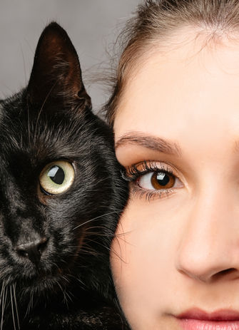 Extreme Nahaufnahme von einem Auge einer Frau und einem Auge einer schwarzen Katze, die Wange an Wange stehen. Beide blicken direkt in die Kamera.