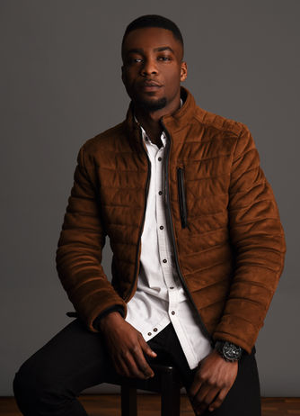 Junger Mann mit brauner Steppjacke und weißem Hemd sitzt auf einem Hocker und schaut ernst in die Kamera.