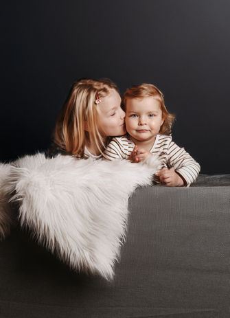 Älteres Kind gibt dem kleineren Kind einen Kuss auf die Wange, beide lehnen auf einem Sofa.