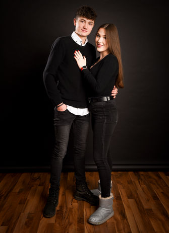 Ein junges Paar steht eng umschlungen vor schwarzem Hintergrund. Beide tragen schwarze Kleidung. Die Frau legt die Hand auf die Brust des Mannes und lächelt leicht.