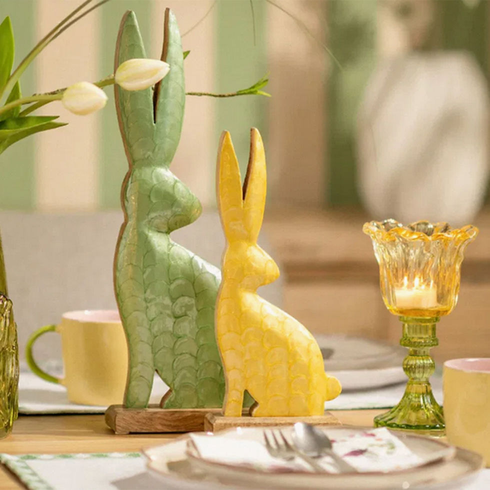 Miniaturbild: Frühlingshafte Tischdekoration mit zwei Hasenfiguren in Grün und Gelb, daneben ein tulpenförmiges Glaswindlicht
