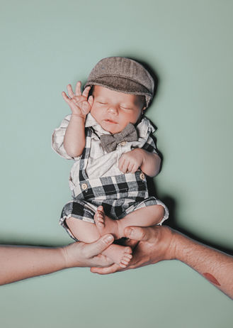 Baby in kariertem Outfit mit Hosenträgern und Schiebermütze wird von zwei Händen gehalten, hebt im Schlaf die rechte Hand.