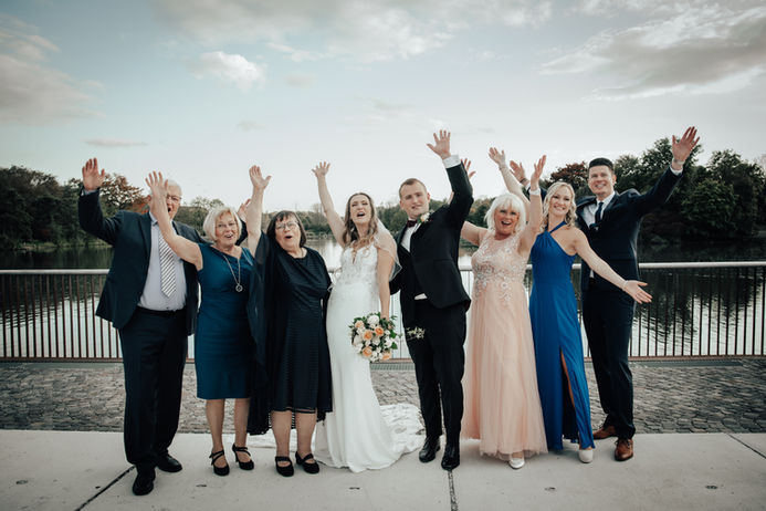 Das Brautpaar steht inmitten einer Gruppe älterer und jüngerer Gäste. Alle winken fröhlich mit erhobenen Armen in die Kamera.