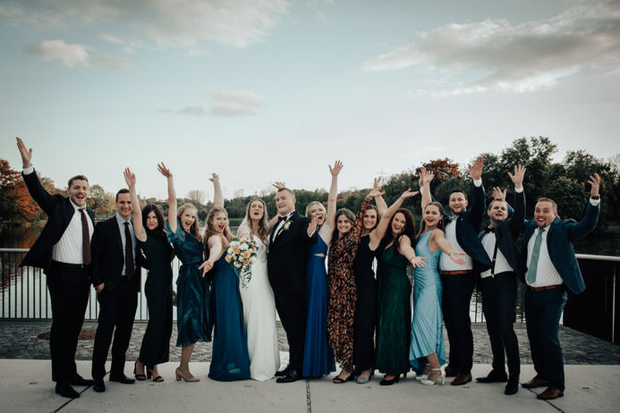 Eine große Gruppe von Hochzeitsgästen versammelt sich um das Brautpaar. Alle lachen, heben die Arme und feiern gemeinsam auf einer Seepromenade.