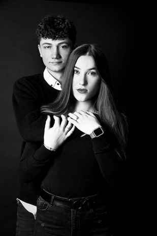 Ein junges Paar vor einem schwarzem Hintergrund. Beide tragen schwarze Oberteile. Der Mann steht hinter der Frau, beide blicken ernst in die Kamera. Der Mann hat einen Arm um den Oberkörper der Frau gelegt, die Frau greift mit bveiden Händen den Arm des Mannes.