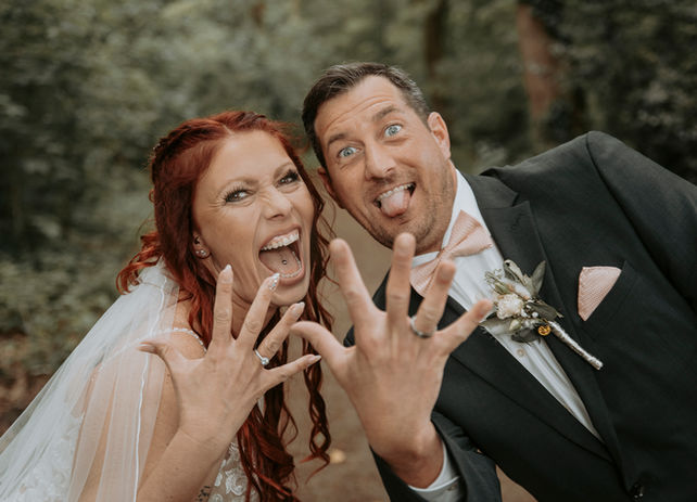 Fröhliches Hochzeitspaar mit viel Humor – die Braut mit leuchtend roten Haaren und der Bräutigam in elegantem Anzug strecken lachend die Zunge heraus und präsentieren stolz ihre Eheringe. Ein modernes, unkonventionelles Hochzeitsfoto voller Lebensfreude!"