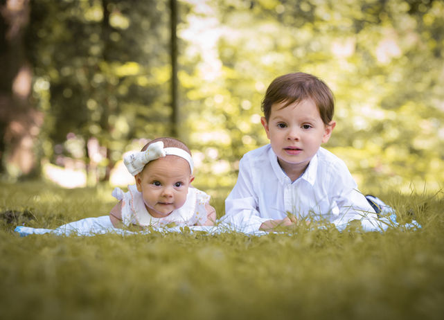 Ein Baby und ein Kleinkind liegen nebeneinander auf einer Decke im Gras. Beide schauen aufmerksam nach vorne, beide tragen weisse Kleidung und das Baby trägt ein Haarreifen mit weisser Schleife.