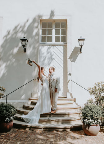 Ein strahlendes Brautpaar steht auf einer eleganten Treppe vor einer historischen, weiß getünchten Fassade mit klassischer Tür und Laternen. Die Braut hebt jubelnd ihren Brautstrauß in die Luft, während sie ihren frisch angetrauten Ehemann verliebt anlächelt. Der Bräutigam, in einem stilvollen cremefarbenen Anzug, erwidert ihren Blick voller Freude. Die Szene wird von sanftem Sonnenlicht durchzogen, das romantische Schattenspiele auf die Wand zaubert und die Leichtigkeit dieses besonderen Moments unterstreicht.